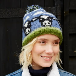 Pachamama Knitwear Dairy Cow Bobble Beanie Hat features blue & green background with knitted Friesian cow heads, 100% Wool, fleece lined